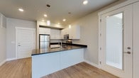 This modern single-floor residence offers a thoughtfully designed open-concept layout that seamlessly connects the kitchen and living area. The kitchen is outfitted with crisp white shaker cabinetry, dark countertops, a peninsula with an integrated sink, stainless steel appliances including a refrigerator, range, and microwave, and stylish pendant lighting that adds a contemporary touch. The living area is a standout feature, anchored by a dramatic curved, floor-to-ceiling glass corner window wall that floods the space with natural light and provides expansive outdoor views. Light wood-look flooring runs throughout the main living areas, adding warmth and cohesion to the design. Two well-proportioned bedrooms are finished with plush carpeting and windows that allow for natural light. The residence includes two bathrooms, each featuring white cabinetry, light wood-look flooring, and a bathtub with shower combination. The primary bathroom is distinguished by a wide vanity with a large framed mirror and bright vanity lighting. Throughout the unit, neutral tones, clean lines, and recessed lighting contribute to a bright, modern, and move-in-ready ambiance.