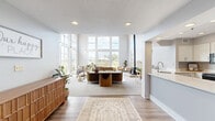 Located at 700 Vista Dr in Gahanna, Ohio, this single-floor community space offers a thoughtfully designed layout ideal for gathering and everyday convenience. The centerpiece is a dramatic living room distinguished by soaring vaulted ceilings supported by striking exposed timber trusses, creating an airy, open atmosphere. Floor-to-ceiling windows flood the space with natural light and frame views of the surrounding landscaping and community grounds, while a stone fireplace anchors one corner, adding warmth and architectural character. The living area is furnished with a tufted sofa, mid-century accent chairs, and lush indoor plants that complement the bright, neutral color palette throughout. A mezzanine-level overlook with a white balustrade railing adds vertical interest and visual depth to the room. Adjacent to the main living area, a kitchenette with light cabinetry provides functional support for events and gatherings. The hallways connect seamlessly to a well-equipped laundry facility featuring multiple front-load washers and dryers set against clean gray walls with tile flooring — practical and efficiently arranged. Throughout the space, recessed lighting, plush carpeting, and consistent soft-toned finishes contribute to a welcoming, polished ambiance suited for a residential community setting.