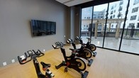 This single-level spin studio is purpose-built for cycling workouts, offering a focused and well-equipped fitness environment. The room features a row of Matrix stationary bikes arranged to face a large wall-mounted flat-screen display, ideal for guided classes or virtual cycling programs. The flooring is a warm-toned hardwood, providing a clean and durable surface suited to high-intensity use.

Neutral gray walls contribute to a focused, distraction-free atmosphere, while a floor-to-ceiling glass window along one wall floods the room with natural light and frames views of the surrounding urban streetscape. This generous glazing creates an open, airy feel despite the room's dedicated single-purpose layout.

The studio's design balances functional simplicity with modern aesthetics, making it a practical space for both solo rides and group cycling sessions. The combination of professional-grade equipment, natural light, and clean finishes gives the studio a polished, gym-quality environment well-suited for daily training.