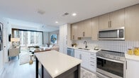 This thoughtfully designed one-bedroom, one-bathroom unit offers a contemporary open-concept layout that maximizes its efficient footprint. The combined kitchen and living area forms the heart of the home, anchored by a striking two-tone kitchen featuring white lower cabinetry, light wood upper cabinets, a vertical subway tile backsplash, and a full suite of stainless steel appliances including a gas range, dishwasher, microwave, and refrigerator. A freestanding island with a quartz countertop provides additional prep space and casual dining. The living area flows naturally from the kitchen and is bathed in natural light from large floor-to-ceiling windows that frame urban views. The bedroom is a serene retreat, highlighted by a textured dark accent wall, a dedicated walk-in closet, and expansive windows that continue the theme of connectivity with the outdoors. The bathroom is finished with a light wood vanity, matte black fixtures, large-format dark tile flooring, a white quartz countertop, and a glass-enclosed walk-in shower. Light wood-style flooring runs throughout the main living areas, tying the space together with a warm, cohesive aesthetic.