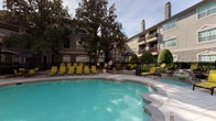 This outdoor pool area is a well-appointed amenity space designed for relaxation and recreation. The centerpiece is a generously sized freeform swimming pool with crystal-clear turquoise water, complemented by an adjacent spa. The surrounding sundeck is finished with light-toned pavers and is furnished with multiple yellow-cushioned chaise lounges, offering ample space for sunbathing and leisure. Mature trees provide natural shade throughout the courtyard, adding a lush, park-like atmosphere to the setting. Additional seating areas with patio furniture are positioned around the perimeter, creating inviting spots for socializing. Neatly trimmed topiaries and potted flowering plants contribute to the manicured landscaping. The courtyard is enclosed by a multi-story building with balconies overlooking the pool, providing a sense of privacy while adding an elegant architectural backdrop. Access to the pool area is provided through a covered concrete corridor, which connects the interior of the building to the outdoor amenity space. The corridor features textured stucco walls and ceiling-mounted lighting, offering a functional and well-lit passageway. Together, these elements create a cohesive and attractive outdoor retreat suitable for year-round enjoyment.