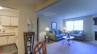 This single-floor residence offers a thoughtfully designed layout that emphasizes openness and comfort. The living area serves as the heart of the home, featuring soft, wall-to-wall carpeting that adds warmth underfoot and a neutral color palette of muted yellows and cool grays that creates a calm, inviting ambiance. A large window floods the space with natural light, brightening the room and enhancing its airy feel. The living room provides generous space for seating and relaxation, with a clear sightline that connects seamlessly to the adjoining dining area. A decorative display shelf near the entryway adds a touch of character and serves as an elegant transition point between the two spaces. Subtle architectural detailing, including a soffit overhead, defines the boundary between the dining and living areas without interrupting the open flow. Recessed lighting supplements the natural light, ensuring the interior remains well-lit throughout the day. The overall design strikes a balance between functionality and comfort, making it well-suited for both everyday living and casual entertaining. The cohesive use of neutral tones throughout ties each area of the home together in a unified, harmonious aesthetic.