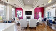 Located at 1320 South Val Vista Drive in Mesa, Arizona, this single-floor office space makes a striking first impression with its bold interior design and thoughtfully curated layout. The space is defined by its dramatic magenta accent walls, which contrast beautifully against a crisp white coffered ceiling and warm hardwood flooring, creating an energetic yet polished atmosphere.

A central focal point is the elegant marble fireplace surround, above which a wall-mounted flat-screen TV is positioned, flanked by the vivid accent walls and decorative greenery. A seating arrangement of white wingback chairs and accent sofas with colorful throw pillows creates a welcoming lounge area ideal for collaboration or reception.

Large windows along the perimeter flood the interior with natural light and offer views of the surrounding outdoor landscape, enhanced by decorative patterned valances and flowing curtains. A dedicated workstation area with blue upholstered counter-height chairs lines one wall, while a sleek white countertop bar provides an additional functional surface.

A ceiling fan, recessed lighting, and a glass-paneled entryway with a secondary work area round out the space. The overall ambiance is modern and inviting, blending professional functionality with vibrant residential-inspired design elements throughout.