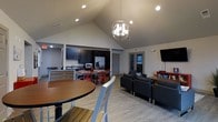 This single-floor space showcases a thoughtfully designed open-concept layout that seamlessly connects the kitchen, dining, and living areas into one cohesive and functional environment. The vaulted ceilings rise dramatically overhead, amplifying the sense of space and airiness throughout the main living area. A striking spherical chandelier serves as a focal point, casting warm light across the light wood-tone flooring that runs throughout.

The kitchen is outfitted with dark cabinetry, stainless steel appliances including a French door refrigerator and microwave, and a functional counter workspace. A kitchen island with seating provides an additional casual dining option. Adjacent to the kitchen, a dedicated dining area features a round table paired with bold red chairs, adding a lively contrast to the otherwise neutral color palette.

The living area is furnished with a set of dark upholstered sofas and chairs arranged around a wall-mounted flat-screen television, creating a comfortable and practical gathering space. A secondary dining or activity table is positioned nearby, offering additional flexibility. Recessed lighting supplements the natural light that filters in through windows near the entry, and neutral gray walls tie the entire space together with a clean, modern aesthetic.