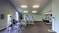 This fitness room offers a functional and well-organized workout space designed to accommodate a range of exercise needs. The room features light gray-green walls and a dark rubber floor, providing a practical and durable surface suited for heavy equipment use. Overhead, multiple flush-mounted fluorescent light fixtures ensure the space is brightly and evenly lit throughout. An arched window on one side of the room allows natural light to filter in, adding a welcoming element to the environment. The equipment layout includes a multi-station cable and weight machine at the center of the room, ideal for a variety of strength training exercises. A squat rack with weight plates is positioned near the window, offering additional free-weight training options. On the cardio side, an elliptical trainer and what appears to be a stationary bike or similar machine are positioned toward the far end of the room. Motivational fitness posters are mounted on the walls, contributing to the gym's functional atmosphere. The single-floor layout ensures easy accessibility, and the generous open floor plan allows for comfortable movement between stations.