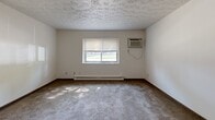 This single-floor unit at 4833 Westchester Dr in Youngstown, Ohio offers a practical and straightforward layout across several well-defined living spaces. The entryway welcomes you through a solid dark wood door, transitioning from updated wood-look vinyl plank flooring into the main living areas. The living space features neutral white walls, textured ceilings, and an abundance of natural light that filters through multiple windows throughout the unit. Several carpeted rooms serve as comfortable bedrooms, each equipped with wall-mounted air conditioning units, baseboard heating, and windows overlooking the surrounding grounds — providing year-round climate control. The galley-style kitchen is efficiently designed with warm wood cabinetry, white laminate countertops, a double sink, a dishwasher, a coil-burner electric range, and a full-size refrigerator, all set on matching vinyl plank flooring. The full bathroom includes a wood-framed vanity mirror with built-in lighting, oak cabinetry, a white countertop sink, and a combined tub-shower unit. Throughout the space, consistent wood-toned trim and neutral finishes create a cohesive, functional aesthetic well-suited for residential living.