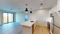 This single-floor residence features a thoughtfully designed open-concept layout with a seamless flow between the kitchen and living areas. The kitchen serves as the centerpiece, anchored by a large island with a white quartz countertop and an undermount sink, illuminated by two striking dark pendant lights. White shaker-style cabinetry lines the walls, complemented by stainless steel appliances including a refrigerator, range, and microwave. Light hardwood flooring extends throughout the main living area, adding warmth to the bright, neutral palette.

The living area offers generous proportions and access to an outdoor balcony through large sliding glass doors, allowing abundant natural light and outdoor views. The bedrooms feature soft carpeting underfoot and large multi-pane windows that frame tree-lined scenery. The primary bathroom is equipped with a wide dual-sink vanity topped with white quartz counters, white shaker cabinets, and large mirrors with modern strip lighting above each sink. A secondary full bathroom includes a tub-shower combination, single vanity, and clean tile flooring. The overall design emphasizes a modern, minimalist aesthetic with consistent finishes throughout.