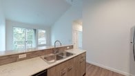 This single-floor unit showcases a thoughtfully designed open-concept layout with vaulted ceilings that lend a sense of generous vertical space to the living and kitchen areas. The kitchen is well-equipped with stainless steel appliances, including an electric range, over-the-range microwave, and refrigerator. Two-toned cabinetry pairs warm wood-grain lower cabinets with crisp white uppers, complemented by light quartz countertops, a subway tile backsplash, a double stainless steel sink, and a pantry accessible through double doors. The living area features warm laminate hardwood flooring and opens directly to a private balcony through sliding glass doors, offering outdoor views of surrounding greenery. The bedroom is carpeted for comfort and includes a sliding-door closet with built-in hanging storage. The bathroom is fitted with a full soaking tub and shower, a wide vanity with ample drawer storage, a large mirror, and wood-look flooring. A stacked washer and dryer unit is conveniently integrated near the bathroom. Throughout the unit, clean white walls, recessed lighting, and consistent modern hardware contribute to a cohesive, contemporary aesthetic.