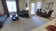 This single-floor space offers a comfortable and functional open-concept layout that seamlessly connects the living and dining areas. The living room is carpeted and thoughtfully arranged, featuring a tufted sofa, a round coffee table, and a side table with a classic lamp, creating a warm and inviting atmosphere. Decorative touches, including a wall-mounted candelabra accent piece and rich burgundy drapes, add character and depth to the space. Large windows fitted with white blinds allow an abundance of natural light to fill the room, enhancing the neutral, earthy tones of the walls and flooring. A glass door leads to an outdoor area, further brightening the interior and providing convenient exterior access. Adjacent to the living room, the dining area is furnished with dark wood chairs and a matching table, providing a defined yet open space for meals. The consistent use of dark-toned furniture against the light carpet and walls creates a cohesive, balanced aesthetic throughout. Overall, this space strikes a comfortable balance between casual everyday living and a welcoming environment suitable for entertaining.