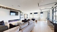 Located at 1050 Slater Rd in Durham, NC, this single-floor amenity space is designed with a contemporary aesthetic that blends bold design elements with functional, inviting areas for gathering and recreation. The lounge area serves as the centerpiece of the space, anchored by a dramatic floor-to-ceiling accent wall clad in dark textured tile, which frames a wall-mounted television and a sleek linear electric fireplace with color-changing flames. Floating shelves and plush seating, including a large sectional sofa and deep lounge chairs arranged over an abstract area rug, create an atmosphere that is both stylish and comfortable. Light wood-look flooring runs throughout, offering a warm contrast to the monochromatic color palette. The kitchen is a standout feature, boasting striking black-and-white vertical tile work along the backsplash wall, bold veined stone countertops, white cabinetry with matte black hardware, stainless steel appliances, and a dark island with pendant lighting overhead. Large windows flood the kitchen with natural light and offer views of the surrounding greenery. The dining area accommodates a large group at a substantial dark table beneath an artistic chandelier, with floor-to-ceiling glass partition doors opening to an adjacent lounge or outdoor area, enhancing the sense of openness and connectivity throughout the space.