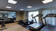 This single-level fitness center offers a practical and well-organized workout space designed to accommodate a range of cardio-focused exercise needs. The room features a selection of modern equipment, including treadmills, elliptical trainers, and a rowing machine, all arranged to maximize the available floor area. The machines are positioned on protective dark rubber matting, which contrasts with the patterned carpeted flooring surrounding it. Natural light enters through vertical-blind-covered windows along one wall, lending a bright, open feel to the space. The ceiling features a drop-tile grid with recessed fluorescent panel lighting, providing consistent, even illumination throughout. A large wall-mounted mirror along one side of the room visually expands the space and allows users to monitor their form during workouts. The neutral wall tones and understated décor keep the environment clean and functional. Overall, the space presents a straightforward, no-frills fitness facility suited for general cardiovascular training.
