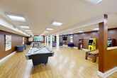 This generously sized single-floor facility is thoughtfully designed as a multifunctional recreational and social space. The interior features warm brown accent walls paired with white structural columns and light hardwood flooring throughout, creating a welcoming and energetic ambiance. Flush-panel ceiling light fixtures provide consistent, even illumination across the open floor plan.

The game room area is a standout highlight, equipped with a full-size pool table, a ping-pong table, a shuffleboard table, a skee-ball lane, and two classic arcade cabinets — an NBA arcade game and a Pac-Man machine — offering a wide variety of entertainment options. Natural light filters in through multiple windows along one wall, enhancing the lively atmosphere.

The lounge area features multiple seating arrangements, including sofas, armchairs, and a round dining table with chairs, providing comfortable spaces for relaxation and conversation. A wall-mounted flat-screen TV with a console table serves as a media focal point. Colorful abstract artwork is displayed throughout, adding visual interest to the rich brown walls. A bar-height table with stools offers an additional casual seating option. The space connects through a hallway, completing this well-rounded, amenity-rich facility.