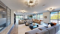 This sophisticated single-floor amenity space is designed to impress, blending contemporary style with comfort in an open, flowing layout. The main lounge area is anchored by multiple large sectional sofas arranged around low-profile coffee tables, creating generous gathering zones ideal for socializing or relaxing. Overhead, a series of striking gold sputnik-style chandeliers serve as dramatic focal points, casting warm, ambient light throughout the space.

A bold stone-clad fireplace commands attention along one wall, adding texture and a sense of warmth to the modern interior. The herringbone-patterned wood flooring runs throughout, lending an elegant, cohesive quality underfoot. Floor-to-ceiling windows and expansive glass panels line the perimeter, flooding the space with natural light and offering open views of the surrounding greenery and outdoor areas.

A dedicated game and recreation area features a shuffleboard table alongside a bistro-height table with seating, positioned beside large windows that frame outdoor garden views. Throughout the space, carefully curated artwork, lush indoor plants, and refined furnishings in warm neutral tones complete the upscale atmosphere. Access to an outdoor terrace is provided through full-height glass doors, seamlessly connecting the interior to the exterior environment.
