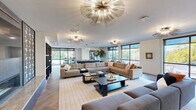 This sophisticated single-floor amenity space is designed to impress, blending contemporary style with comfort in an open, flowing layout. The main lounge area is anchored by multiple large sectional sofas arranged around low-profile coffee tables, creating generous gathering zones ideal for socializing or relaxing. Overhead, a series of striking gold sputnik-style chandeliers serve as dramatic focal points, casting warm, ambient light throughout the space.

A bold stone-clad fireplace commands attention along one wall, adding texture and a sense of warmth to the modern interior. The herringbone-patterned wood flooring runs throughout, lending an elegant, cohesive quality underfoot. Floor-to-ceiling windows and expansive glass panels line the perimeter, flooding the space with natural light and offering open views of the surrounding greenery and outdoor areas.

A dedicated game and recreation area features a shuffleboard table alongside a bistro-height table with seating, positioned beside large windows that frame outdoor garden views. Throughout the space, carefully curated artwork, lush indoor plants, and refined furnishings in warm neutral tones complete the upscale atmosphere. Access to an outdoor terrace is provided through full-height glass doors, seamlessly connecting the interior to the exterior environment.
