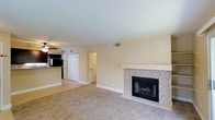 This single-floor space presents a welcoming and functional layout with a clean, neutral aesthetic. The main living area is carpeted in a light beige tone, providing a soft and comfortable underfoot feel that complements the warm, cream-colored walls throughout. A standout feature of the living area is the tile-surround fireplace, finished with a mosaic-style tile pattern in neutral tones and topped with a simple white mantel, serving as a natural focal point in the room. Overhead, a flat white ceiling with a visible HVAC vent contributes to the tidy, well-maintained appearance of the space. Natural light is supplemented by ceiling-mounted fixtures, maintaining a bright and airy atmosphere. Adjacent to the living area, a partial view of the kitchen reveals dark appliances and a raised countertop, suggesting an open or semi-open connection between spaces. Two white paneled doors are visible in the background, indicating access to additional rooms or storage areas, pointing to a practical and organized floor plan. Overall, the space offers a straightforward, move-in-ready environment with comfortable finishes and a cohesive design sensibility.