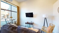 This well-appointed 1-bedroom, 1-bathroom unit in Jonesboro, AR offers a thoughtfully designed single-floor layout that balances modern aesthetics with everyday functionality. The living area is bright and open, anchored by large floor-to-ceiling windows dressed with warm golden curtains that flood the space with natural light. Polished concrete floors run throughout, lending an industrial-chic character to the interiors. A wall-mounted flat-screen TV, hairpin-leg side tables, and a bistro dining set positioned by the windows complete the living space. A wall-mounted mini-split system ensures year-round climate comfort. The kitchen is a standout feature, showcasing bold navy lower cabinets paired with light upper cabinetry, rich butcher block countertops, a matte black faucet, and full appliances including an electric range, dishwasher, and over-the-range microwave. The bedroom offers a serene retreat with crisp white walls, vaulted ceilings, a dark-framed bed with neutral linens, and botanical wall art that adds a calming touch. The bathroom is clean and functional, featuring a tub-shower combination. A private balcony accessible through a glass-paneled door extends the living space outdoors, providing a cozy spot to enjoy views of the surrounding streetscape.