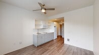 Located at 2211 SW Fourth Ave in Portland, Oregon, this efficiently designed studio unit offers a well-organized single-floor layout that maximizes every square foot. The open living area features warm wood-style laminate flooring throughout and crisp white walls that create a clean, modern aesthetic. A ceiling fan with a built-in light fixture anchors the main living space, which is filled with natural light from a pair of large windows fitted with horizontal blinds, offering pleasant views of surrounding greenery.

The kitchen is thoughtfully appointed along one wall, featuring light gray cabinetry with modern hardware, a white quartz countertop, a single-basin sink, an electric two-burner cooktop, an over-the-range microwave, a refrigerator, and open upper shelving for additional storage. The layout keeps the kitchen easily accessible without encroaching on the living area.

The bathroom is finished with the same wood-style flooring and includes a walk-in shower, a toilet, and stainless steel grab bars and towel bars. A dedicated electrical panel is conveniently recessed into the wall near the entryway. The unit also includes a floor-level heating vent and a smoke detector, ensuring comfort and safety throughout.