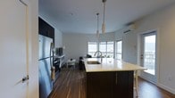 Located at 36 Pine St in Poughkeepsie, NY, this one-bedroom, one-bathroom residence offers a thoughtfully designed single-floor layout that maximizes both functionality and style. The open-concept living and kitchen area serves as the heart of the home, creating a seamless flow between spaces. The kitchen is outfitted with sleek dark cabinetry, stainless steel appliances including a full-size refrigerator and range, and a spacious center island with a light-toned countertop and integrated sink — ideal for both meal preparation and casual dining. Pendant lighting above the island adds a refined touch to the space. Rich, dark hardwood flooring runs throughout, grounding the interior with warmth and contrast against the light-toned walls and ceilings. The living area transitions naturally from the kitchen, offering a comfortable and airy environment. A wall-mounted ductless HVAC unit ensures year-round climate comfort. Large windows along the far wall flood the space with natural light and provide open views, enhancing the sense of spaciousness. The overall aesthetic is clean, modern, and functional — a well-appointed residence suited for contemporary urban living in the heart of Poughkeepsie.
