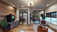 Located on N Pecan Pkwy in Justin, Texas, this single-story clubhouse facility presents a thoughtfully designed interior that blends modern industrial aesthetics with warm, rustic touches. Rich hardwood flooring flows throughout the space, complemented by reclaimed wood accent walls that add texture and character to key areas.

The main reception and lounge area serves as the heart of the facility, anchored by a striking sputnik chandelier with gold and wood accents. Glass-partitioned offices with black steel frames line the space, offering transparency while maintaining functional separation. Each private office is furnished with desks and seating, ideal for individual meetings or focused work.

A welcoming lounge area features a full-height stacked stone fireplace with a mounted flat-screen television, framed by French doors that open to the exterior. Overhead, a rustic wagon-wheel chandelier with Edison bulbs adds warmth and ambiance. Comfortable seating arrangements invite relaxation throughout.

The kitchen is well-appointed with dark cabinetry, marble-style countertops, a peninsula with bar seating, open wood shelving, decorative encaustic tile backsplash, pendant lighting, and stainless steel appliances. A dedicated fitness room, accessed through a glass sliding door set into a reclaimed wood wall, houses exercise equipment for resident use.