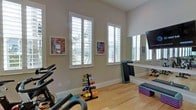 This dedicated fitness space is thoughtfully designed to support a comprehensive workout routine. The room features light hardwood flooring that adds warmth underfoot, while the neutral gray walls create a clean, focused environment. Multiple windows fitted with classic plantation shutters allow generous natural light to fill the space, contributing to an open and energizing atmosphere. A large wall-mounted mirror runs along one side of the room, visually expanding the space and allowing for form-checking during exercise. A wall-mounted television screen provides entertainment or instructional content during workouts. The space is well-equipped with a range of fitness equipment, including stationary cycling bikes and a tiered dumbbell rack stocked with color-coded free weights in various sizes. Additional accessories such as aerobic step platforms, resistance mats, and other training tools are neatly organized throughout the room. Motivational framed artwork adorns the walls, adding a personal and uplifting touch to the environment. The layout is practical and spacious, allowing for both cardio and strength training activities. Overall, this fitness room offers a fully functional, inviting, and well-lit setting ideal for daily exercise.