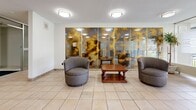 The lobby at 345 Sentinel Road in Toronto, Ontario, offers a well-appointed and functional entry experience for residents and visitors alike. The space is organized across two distinct zones seamlessly connected by light-toned ceramic tile flooring throughout.

The entrance vestibule makes an immediate impression with its floor-to-ceiling glass curtain wall system, flooding the space with natural light while providing clear sightlines to the exterior. The building's address is prominently displayed on the glazing, and a set of glass doors with frosted banding provides a secure yet inviting transition point. A seating area just inside features a pair of rounded grey upholstered chairs arranged around a classic wooden coffee table, set against a striking decorative mirror wall with an amber-toned patina finish that adds warmth and visual interest.

A standout architectural element is the ornate dark wood reception console, featuring elaborate carved columns, crown moulding, and decorative detailing — a classic piece that lends the lobby a distinguished, traditional character amid its otherwise clean, contemporary surroundings.

The inner lobby leads directly to a dedicated elevator hall, where two stainless steel elevator doors are set against warm stone-toned wall cladding. Flush ceiling light fixtures provide consistent, even illumination throughout, while grey runner mats at key transition points add practicality. The overall ambiance is clean, bright, and professional.