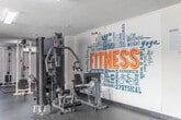 Located at 1867 Bloor St in Mississauga, Ontario, this fitness centre offers a well-organized and comprehensively equipped workout space spread across a single floor. The room features durable rubber flooring throughout, providing both safety and comfort during exercise. Overhead fluorescent lighting ensures the space is brightly and evenly lit, while a small window admits a touch of natural light.

The facility is thoughtfully divided into functional zones. The cardio area houses a treadmill and an elliptical trainer, ideal for aerobic workouts. The free weights section includes a full dumbbell rack with an assortment of weights, a flat bench, and a barbell with weight plates. A dedicated stretching and floor exercise area features protective rubber mats. A prominent rack holds an extensive collection of medicine balls, resistance bands, and stability balls of various sizes, supporting functional and core training.

Strength training is well-served by a multi-station cable machine and a power rack with a barbell and plates, as well as a captain's chair for core work. A large wall mirror spans one side of the room, enhancing the sense of space and allowing users to monitor their form. The standout design feature is a bold, large-scale motivational mural on one wall, displaying fitness-themed words and phrases in vibrant typography, energizing the atmosphere of the room.