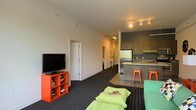 This single-floor space offers a contemporary, open-concept design that blends functionality with bold, energetic style. The living area is anchored by a sleek flat-screen television mounted on a striking orange shelving unit, which serves as a focal point against the clean white walls. Lime green seating and ottoman accents bring a lively, youthful energy to the space, complemented by dark, patterned carpeting that adds depth and texture underfoot.

The layout flows naturally into the kitchen, which features a modern peninsula with gray cabinetry and warm wood-toned countertops. Orange metal barstools line the counter, maintaining the space's vibrant color palette. The kitchen is equipped with upper cabinetry with under-cabinet lighting and includes full-size appliances, with tile flooring that transitions cleanly from the carpeted living area.

Track lighting runs along the ceiling in the kitchen area, providing practical illumination, while recessed lighting ensures the living space remains well-lit throughout. A hallway visible from the main room leads toward additional rooms, suggesting a thoughtfully connected floor plan. The overall ambiance is modern and dynamic, with a carefully curated use of contrasting colors and contemporary finishes that create an inviting, lively atmosphere throughout.