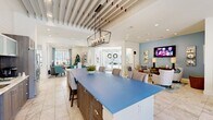 Located at 14300 Sapphire Park Ln in Midlothian, Virginia, this single-story community clubhouse offers a thoughtfully designed, open-concept gathering space with a bright and inviting atmosphere throughout. The interior is anchored by a striking oversized kitchen island with a bold blue countertop, complemented by dark wood cabinetry, glass-front upper cabinets, a mosaic tile backsplash, and stainless steel appliances. An eye-catching lantern-style chandelier hangs above, while a distinctive slatted ceiling detail adds architectural character to the space. Large format marble-look tile flooring runs throughout, enhancing the sense of spaciousness and light. Adjacent to the kitchen, a casual dining nook features a round white table and teal upholstered chairs beside floor-to-ceiling windows and a glass door that opens directly to the outdoor pool area, flooding the space with natural light and outdoor views. A dedicated lounge area provides comfortable seating arranged around a wall-mounted flat-screen television set against a soft blue accent wall, creating an ideal space for relaxed social gatherings. The welcoming entry foyer features soaring ceilings and expansive glass double doors. The overall design palette of whites, blues, and warm wood tones establishes a fresh, coastal-inspired ambiance throughout.