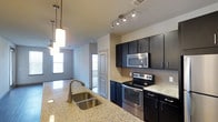 This single-story Texas residence showcases a thoughtfully designed interior with a strong emphasis on modern aesthetics. The kitchen serves as a central highlight, featuring rich dark cabinetry that extends floor-to-ceiling, offering generous storage throughout. Light-toned granite countertops provide a striking contrast against the dark cabinet finish, lending the space a polished, contemporary look. A stainless steel double-basin sink anchors the kitchen island, accompanied by elegant pendant lighting that hangs in a cascading arrangement, adding both function and visual interest. Stainless steel appliances, including an electric range, over-the-range microwave, and dishwasher, contribute to the kitchen's cohesive, upscale appearance. A classic white subway tile backsplash adds texture and a timeless touch behind the range area. Track lighting along the ceiling ensures thorough illumination across the workspace. A glimpse beyond the kitchen reveals a connected living area bathed in natural light from nearby windows, enhancing the open, airy feel of the single-floor layout. The overall ambiance is clean, modern, and well-appointed, suited for everyday living and entertaining alike.