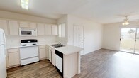 This single-floor residence offers a clean, open-concept layout that seamlessly connects the kitchen and living areas, creating a sense of spaciousness and easy flow throughout the home. The kitchen is thoughtfully designed with light-toned cabinetry that extends to the ceiling, providing ample storage space. A peninsula with a built-in sink and breakfast bar serves as the centerpiece, offering both workspace and a casual dining option. White appliances, including an electric range, built-in microwave, and dishwasher, are neatly integrated into the layout. A window above the sink allows natural light to brighten the workspace. The kitchen transitions naturally into the open living area, which continues the warm wood-look flooring found throughout the space. This consistent flooring choice unifies the rooms and adds a modern, welcoming touch. The neutral white walls and bright, airy palette throughout create a versatile backdrop well-suited for a variety of interior styles. The living area benefits from generous open space, and a paneled door provides access to an adjacent room or entry point, adding to the home's practical and connected floor plan.