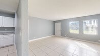 Located at 1001 36th St in West Palm Beach, Florida, this single-story home presents a clean, well-maintained interior with a cohesive and functional layout. Throughout the home, large-format white tile flooring reflects natural light and creates a bright, airy atmosphere. The walls are painted in a soft, calming blue-gray tone, complemented by crisp white baseboards that add a polished finish to each room.

The main living area is generously proportioned, offering an open and flexible space ideal for a variety of configurations. Natural light filters in through windows fitted with vertical blinds, adding to the home's inviting ambiance. The layout flows seamlessly from the living area into additional rooms, including what appears to be a secondary living or dining space.

The kitchen features white shaker-style cabinetry, granite countertops with subtle speckled detailing, a stainless steel sink, and a full-size refrigerator. An adjacent closet provides convenient pantry or storage access.

The bathroom is thoughtfully appointed with marble-look tile surrounding the tub and shower, complemented by coordinating floor tile, a white vanity with granite countertop, and a gold-framed mirror that adds a warm accent.

Additional rooms include a bedroom-sized space with the same consistent tile flooring and central air supply vent, maintaining the home's uniform, well-kept character throughout.