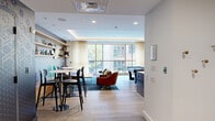 Located at 65 Brainerd Road in Boston's Allston neighborhood, this thoughtfully designed single-floor lounge space offers a vibrant and versatile environment for socializing and recreation. The open layout is anchored by light hardwood flooring and a tray ceiling with warm ambient LED cove lighting, lending the room a polished, contemporary feel.

The space is organized into distinct functional zones. A casual lounge area features a bold, patterned navy accent wall adorned with geometric wallcovering and open wood shelving displaying curated décor. Leather armchairs, colorful ottoman stools, and a live-edge coffee table create an inviting gathering spot centered around a wall-mounted flat-screen TV.

Adjacent to this is a billiards area with a full-size pool table, complemented by framed artwork and a built-in counter with bar-height seating. A high-top dining area near the entrance offers additional seating with bistro-style tables and modern stools.

Large floor-to-ceiling windows along one wall flood the space with natural light and provide views of the surrounding neighborhood. Hexagonal mirror wall art and warm wood cabinetry add decorative detail near the entry corridor, which features stylish patterned double doors. The overall ambiance is energetic yet comfortable, balancing modern design elements with functional amenities suited for a shared community setting.