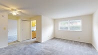 This single-floor unit offers a straightforward and functional layout across several well-defined spaces. The living area is generously sized, featuring neutral wall tones and soft grey wall-to-wall carpeting that extends throughout the main living spaces. A large window fitted with horizontal blinds allows natural light to fill the room while offering outdoor views of surrounding greenery. A sliding closet door provides convenient built-in storage adjacent to the living area. The entry door opens directly into this expansive room, contributing to an open and airy feel.

The galley-style kitchen is efficiently arranged, with warm honey-toned wood cabinetry lining both walls and providing ample storage above and below the counters. Blue-toned laminate countertops complement the cabinetry, and the space includes a white gas range and a stainless steel sink. A window at the end of the kitchen brings in additional natural light.

The bedroom is a comfortable, well-proportioned room with matching grey carpeting and its own window with blinds. A ceiling fan with an integrated light fixture provides both air circulation and overhead lighting.

The bathroom features a full soaking tub with a tiled surround in clean white, finished with chrome fixtures, creating a simple and practical bathing space.