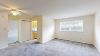 This single-floor unit offers a straightforward and functional layout across several well-defined spaces. The living area is generously sized, featuring neutral wall tones and soft grey wall-to-wall carpeting that extends throughout the main living spaces. A large window fitted with horizontal blinds allows natural light to fill the room while offering outdoor views of surrounding greenery. A sliding closet door provides convenient built-in storage adjacent to the living area. The entry door opens directly into this expansive room, contributing to an open and airy feel.

The galley-style kitchen is efficiently arranged, with warm honey-toned wood cabinetry lining both walls and providing ample storage above and below the counters. Blue-toned laminate countertops complement the cabinetry, and the space includes a white gas range and a stainless steel sink. A window at the end of the kitchen brings in additional natural light.

The bedroom is a comfortable, well-proportioned room with matching grey carpeting and its own window with blinds. A ceiling fan with an integrated light fixture provides both air circulation and overhead lighting.

The bathroom features a full soaking tub with a tiled surround in clean white, finished with chrome fixtures, creating a simple and practical bathing space.