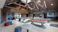 This expansive single-floor community lounge space makes an immediate impression with its dramatic vaulted ceiling, where exposed wooden beams and metal hardware create a bold industrial aesthetic. The raw texture of exposed brick walls contrasts beautifully with sleek modern finishes throughout the space, lending it a refined yet relaxed atmosphere.

The open-plan layout is divided into distinct activity zones designed for socializing and recreation. A central lounge area features a generous sectional sofa and modern cube seating arranged on a teal area rug, ideal for casual gatherings. Adjacent to this is a pool table with a striking orange felt surface, serving as a natural entertainment focal point. High-top round tables with modern stools provide additional seating throughout.

Toward the back, a bar-style counter area with pendant lighting and a wall-mounted flat-screen TV creates an inviting space for watching sports or casual conversation. Sleek linear pendant lighting and halo-style fixtures suspended from the ceiling add a contemporary touch to the rustic framework above.

Large windows allow natural light to filter into the space, enhancing the open and energetic feel. The polished tile flooring ties the various zones together seamlessly, making this a well-designed, versatile space built for community engagement and entertainment.
