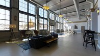 This impressive single-floor space showcases a bold industrial aesthetic, characterized by soaring ceilings with exposed ductwork and structural elements that speak to its warehouse or factory origins. Oversized, multi-pane steel-framed windows line the walls, stretching from floor to ceiling and flooding the interior with abundant natural light while offering views of the surrounding neighborhood. The open-plan layout is anchored by polished concrete floors that reflect the daylight streaming in, enhancing the airy, expansive feel of the space. The main area features a comfortable lounge arrangement with tufted navy seating, a rustic wooden bench, and layered rugs, creating an inviting gathering zone within the grand volume. Large pendant lights with warm globe shades hang from above, complementing the industrial architecture with a refined touch. Toward the rear, additional functional zones are visible, including workspaces and equipment areas, suggesting a versatile, multi-use environment. Structural columns punctuate the space at regular intervals, reinforcing the building's heritage while seamlessly integrating with the contemporary fit-out. The combination of raw architectural elements, generous proportions, and thoughtful furnishings creates a dynamic and inspiring atmosphere throughout.
