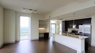 This contemporary single-floor residence showcases a thoughtfully designed open-concept layout that seamlessly blends living, dining, and kitchen spaces. Rich dark hardwood flooring extends throughout, anchoring the space with warmth and sophistication. Floor-to-ceiling windows flood the interior with abundant natural light while offering sweeping elevated views of the surrounding cityscape and greenery, creating an airy, expansive atmosphere. The modern kitchen features dark espresso cabinetry, granite countertops, stainless steel appliances, and a peninsula with bar seating that opens to the main living area — ideal for both everyday living and entertaining. Pendant lighting and track lighting add functional yet stylish illumination throughout. A built-in desk nook with under-cabinet lighting provides a dedicated workspace that integrates neatly into the living area without disrupting the open flow. The clean white walls and neutral palette serve as an elegant backdrop, allowing the architectural details and high-end finishes to take center stage. Overall, the space exudes a polished, urban aesthetic with a bright, welcoming ambiance well-suited for modern living.
