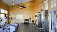 The Willow Creek Resort fitness center offers a thoughtfully designed workout space housed within a single-floor facility. The room features soaring ceilings painted in warm amber tones, creating an energizing yet welcoming atmosphere. A ceiling fan provides comfortable air circulation during workouts, complementing the track lighting mounted above for optimal visibility throughout the space.

The flooring consists of durable dark rubber matting, ideal for supporting heavy equipment and high-impact activity. Cardio equipment, including elliptical machines, is positioned along one side of the room, offering accessible options for aerobic training. A dedicated free weight area features a full rack of dumbbells arranged in ascending order, paired with an adjustable workout bench. On the opposite side, a multi-function cable and weight stack machine provides versatile strength training options.

A mirrored wall enhances the sense of space and allows for form monitoring during exercise. French doors with glass panels allow natural light to filter in, connecting the interior to the broader resort environment. The Willow Creek Resort logo is prominently displayed on the wall, reinforcing the branded, resort-quality experience throughout the space.