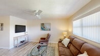 Located in Okeechobee, FL, this well-appointed single-floor, one-bedroom, one-bathroom residence offers a thoughtfully organized layout that maximizes every inch of its comfortable footprint. The open-plan living and dining area serves as the heart of the home, featuring light wood-style flooring, a textured ceiling, and a ceiling fan that enhances the airy Florida ambiance. Natural light filters in through slatted vertical blinds, and the space is anchored by a wall-mounted television and a classic white media console. A glass oval coffee table and plush seating round out the inviting living area. The front entry opens directly into this shared living-dining space, reinforcing the open and connected feel of the home. The kitchen is conveniently positioned off the main living area, while a hallway leads to the generously sized bedroom. The primary bedroom accommodates a full suite of wooden furniture, benefits from its own ceiling fan, and features two closets with louvered doors — offering excellent storage. A window provides natural light and outdoor views. The full bathroom is clean and functional, with white cabinetry, a single vanity, and a shower-tub combination with sliding glass doors. Light, neutral tones are carried consistently throughout the home, creating a cohesive and calm atmosphere.