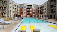 This contemporary amenity space is part of a multi-story residential community distinguished by a mixed-material exterior of brick and horizontal siding. At the heart of the property lies a generously sized outdoor swimming pool, framed by a wide concrete pool deck lined with lounge chairs and umbrellas. The pool area is enclosed on multiple sides by the building's curved and angular wings, creating a semi-private courtyard atmosphere ideal for recreation and leisure.

Inside, the amenity level features a stylish billiards lounge with elevated ceilings and large floor-to-ceiling windows that flood the space with natural light and provide direct views of the pool courtyard. Patterned drapery adds a refined touch to the room's contemporary aesthetic.

Adjacent to the lounge is a well-appointed kitchenette, outfitted with sleek cabinetry, white quartz countertops, a stainless steel sink, and integrated appliances including a refrigerator and microwave. A herringbone-patterned tile backsplash runs behind a wall-mounted flat-screen television, lending the space a polished, modern character. Polished concrete flooring and exposed ceiling beams throughout the interior reinforce an industrial-chic design sensibility. Together, these amenities offer residents a cohesive and versatile space for both casual and social gatherings.