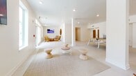 This thoughtfully designed single-floor amenity space combines open-plan recreation with intimate lounge areas, all unified by a clean, modern aesthetic. Large-format white tile flooring anchors the main recreational zone, which centers around a full ping-pong table and is framed by a distinctive slatted wood accent wall that adds warmth and texture to the otherwise crisp white interior. Structural columns divide the expansive open area into naturally defined zones without compromising the sense of spaciousness. Recessed lighting throughout ensures a bright, even atmosphere complemented by generous natural light streaming in through multiple large windows and glass doors that offer views of palm trees outside. A dedicated kitchenette and bar area features warm wood cabinetry, a long white countertop, and bar seating, making it well-suited for social gatherings. A secondary game station, including a foosball table, occupies a corner near the outdoor-facing glass doors. A private TV lounge alcove is furnished with mid-century modern rattan and wood chairs arranged around a woven ottoman, creating an intimate viewing area. The main lounge zone features sculptural wooden stools, a glass coffee table, and designer accent chairs arranged atop a natural fiber area rug, conveying a relaxed yet refined ambiance. A corridor with private rooms provides additional functional space off the main area.