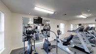 The Hi-Point Apartments Fitness Center is a dedicated, single-floor exercise facility designed to support the health and wellness needs of residents. The room is outfitted with a well-rounded selection of cardio machines, including ellipticals, stationary bikes, and treadmills, providing options for a full range of cardiovascular workouts. Large wall-mounted mirrors span one side of the room, creating a sense of expanded space while allowing users to monitor their form during exercise. A flat-screen television is mounted on the mirrored wall, offering entertainment during workouts. The facility is brightly lit with overhead fluorescent lighting, ensuring a clear and energizing environment. Natural light supplements the interior lighting through windows along the exterior wall, contributing to an open and airy feel. The flooring features a durable, easy-to-clean surface with protective rubber mats positioned beneath the equipment. The walls are finished in a clean, neutral white, giving the space a crisp and functional aesthetic. Informational signage is posted on the walls, providing guidance for facility use. Overall, the fitness center offers a practical and well-equipped space for residents to maintain an active lifestyle without leaving the property.