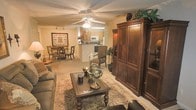 This single-floor residence exudes a classic, refined ambiance through its thoughtfully designed open-concept layout. The living area serves as the heart of the space, anchored by a large, dark-wood entertainment armoire and complemented by a plush seating arrangement set atop a traditional area rug. A substantial wooden coffee table with ornate legs adds a distinguished focal point to the room, while decorative accents — including a globe sculpture and lush greenery — lend character and warmth throughout.

A ceiling fan with an integrated light fixture provides both comfort and illumination, keeping the space well-lit and airy. Crown molding detail along the ceiling adds an elegant architectural touch. The open sightlines connect the living room seamlessly to the dining area, which features a classic round table surrounded by dark wood chairs beneath a track lighting fixture — ideal for intimate gatherings.

The kitchen is positioned just beyond the dining area and features light wood cabinetry, a raised breakfast bar, and white appliances, offering a clean and functional cooking environment. The neutral, warm-toned walls and light flooring tie all areas of the space together cohesively, creating a comfortable and inviting atmosphere throughout.