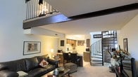 This two-floor loft-style space showcases a thoughtfully designed open-concept layout that maximizes both functionality and visual interest. The main level features a comfortable living area anchored by a leather sofa and a coffee table, seamlessly flowing into a dining area and a well-appointed kitchen. The kitchen is equipped with wood-toned cabinetry, ample counter space, and essential appliances, making it both practical and inviting. Warm, layered lighting throughout the main floor creates a cozy and welcoming ambiance. A striking wrought-iron spiral staircase on one side of the space serves as both a functional element and a design focal point, leading up to the mezzanine level above. The upper level features an open railing with wrought-iron balusters that overlooks the main living area below, adding a sense of vertical depth and openness to the overall space. Textured ceilings and exposed structural beams contribute to the loft's character, while neutral wall tones and carpeted flooring provide a versatile backdrop. The interplay between the two levels creates a dynamic, multi-dimensional living environment well-suited for those who appreciate architectural interest and an efficient use of space.