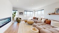 Located on Boulevard de Lucerne in Gatineau, this striking two-floor space delivers a seamless blend of modern architecture and refined design. The main floor commands attention with its dramatic double-height ceiling, flooded with natural light through oversized windows and a large skylight above. Light oak hardwood flooring flows throughout, unifying the spacious open-concept layout.

The kitchen is a standout feature, showcasing warm wood cabinetry paired with crisp white lower cabinets, a clean-lined white waterfall island with bar seating, and sleek pendant lighting. It connects effortlessly to the dining area, which enjoys sweeping panoramic views through floor-to-ceiling sliding doors leading to an outdoor balcony. A circular wood-framed mirror adds an artistic focal point to this bright, airy room.

The main living room impresses with a linear gas fireplace set into a bold white accent wall, surrounded by expansive windows that frame city and landscape views on multiple sides. A second lounge area features a textured vertical wood slat accent wall and direct outdoor access.

A sculptural staircase with dark steel treads and glass railings connects to the upper floor, where an additional living room offers more open space and views. The double-height void visible from the staircase landing reinforces the grandeur of the overall design, creating a truly impressive, light-filled environment.