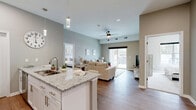 This well-appointed 2-bedroom, 2-bathroom residence in Getzville, NY offers a thoughtfully designed open-concept layout across a single floor. The kitchen serves as a central hub, showcasing white shaker cabinetry, granite countertops, stainless steel appliances, and a spacious island with seating for three — illuminated by stylish pendant lighting. The kitchen flows seamlessly into the living room, which features light hardwood-style flooring, a ceiling fan, and a sliding glass door that invites natural light and outdoor access. Both bedrooms are carpeted and finished in warm neutral tones, providing a calm and restful atmosphere. The generously sized primary bedroom offers ample space for a full furniture arrangement. Each bathroom features granite-topped white vanities and hardwood-style flooring, maintaining a consistent and polished aesthetic throughout. A dedicated laundry room houses a full-size front-loading washer and dryer. Additional storage is provided by a closet. The overall design palette — soft grays, warm beiges, and crisp whites — lends a clean, modern feel that is both functional and welcoming.