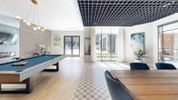 Located at 100 Hamilton St in Bound Brook, NJ, this single-floor amenity lounge is thoughtfully designed to accommodate a variety of social and recreational activities within a generously proportioned open layout. The space is divided into several distinct zones, each with its own character while maintaining a cohesive contemporary aesthetic throughout.

The game and dining area features a striking geometric lattice ceiling in dark matte finish, creating a bold architectural contrast against the bright white walls. Patterned floor tiles define this zone, which includes a billiards table and a dining table surrounded by blue upholstered chairs. A built-in wet bar with teal cabinetry and a wall-mounted television serves as a functional focal point.

Adjacent to this area, a lounge zone offers a warm, residential feel anchored by a sleek linear fireplace set into a large-format stone accent wall, topped by a mounted television and flanked by integrated shelving. Textured carpeting and a large window with outdoor views enhance the comfort of this seating area.

Near the entry, a welcoming seating nook with navy velvet chairs and hardwood flooring provides an inviting transition into the space. Floor-to-ceiling glass doors open to an exterior courtyard, flooding the interior with natural light.