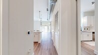 This thoughtfully designed single-floor space blends modern aesthetics with industrial-inspired details to create a bright and welcoming environment. The open-concept living area is anchored by wide-plank hardwood-style flooring that runs throughout, complemented by crisp white walls and abundant natural light streaming in through large floor-to-ceiling sliding glass doors. A ceiling fan and exposed metallic ductwork overhead lend an urban, loft-like character to the living space, while sheer curtain panels frame the outdoor view and soften the room's clean lines.

The galley-style kitchen is efficiently laid out and well-appointed, featuring white shaker cabinetry, classic subway tile backsplash, light countertops, and a full suite of stainless steel appliances including a refrigerator, range, microwave, and dishwasher. Pendant lighting above the kitchen pass-through adds warmth and connects the kitchen to the living area seamlessly.

A hallway leads to additional rooms, lined with louvered-door storage closets that offer generous organizational space. The bathroom features a full tub and shower combination with white subway tile surround, maintaining the clean, minimal aesthetic found throughout the unit. A smart thermostat near the entryway adds a contemporary, tech-forward convenience to the overall living experience.