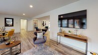 This single-floor communal space is thoughtfully designed to serve as a welcoming gathering hub. The open-concept layout is anchored by light wood-look flooring that flows seamlessly throughout, complemented by neutral white walls and recessed lighting that create a clean, contemporary atmosphere.

The lounge area offers generous seating, including a bold mustard-yellow sofa and accent chairs arranged around a large area rug, with a wall-mounted flat-screen TV providing a media-ready focal point. An aerial view reveals the full extent of the open floor plan, illustrating how the various seating zones connect naturally without feeling crowded.

A dedicated coffee bar serves as a standout feature, showcasing deep navy-blue cabinetry with gold hardware, white subway tile backsplash, white countertops, and a stainless steel under-counter refrigerator — all centered around a fully equipped coffee station.

The dining nook accommodates a round wood table paired with vibrant yellow upholstered chairs, ideal for casual meetings or social gatherings. Large windows with blinds line the perimeter, flooding the interior with natural light, while glass double doors open directly to an outdoor patio and landscaped grounds, enhancing connectivity to the exterior.