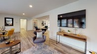 This single-floor communal space is thoughtfully designed to serve as a welcoming gathering hub. The open-concept layout is anchored by light wood-look flooring that flows seamlessly throughout, complemented by neutral white walls and recessed lighting that create a clean, contemporary atmosphere.

The lounge area offers generous seating, including a bold mustard-yellow sofa and accent chairs arranged around a large area rug, with a wall-mounted flat-screen TV providing a media-ready focal point. An aerial view reveals the full extent of the open floor plan, illustrating how the various seating zones connect naturally without feeling crowded.

A dedicated coffee bar serves as a standout feature, showcasing deep navy-blue cabinetry with gold hardware, white subway tile backsplash, white countertops, and a stainless steel under-counter refrigerator — all centered around a fully equipped coffee station.

The dining nook accommodates a round wood table paired with vibrant yellow upholstered chairs, ideal for casual meetings or social gatherings. Large windows with blinds line the perimeter, flooding the interior with natural light, while glass double doors open directly to an outdoor patio and landscaped grounds, enhancing connectivity to the exterior.